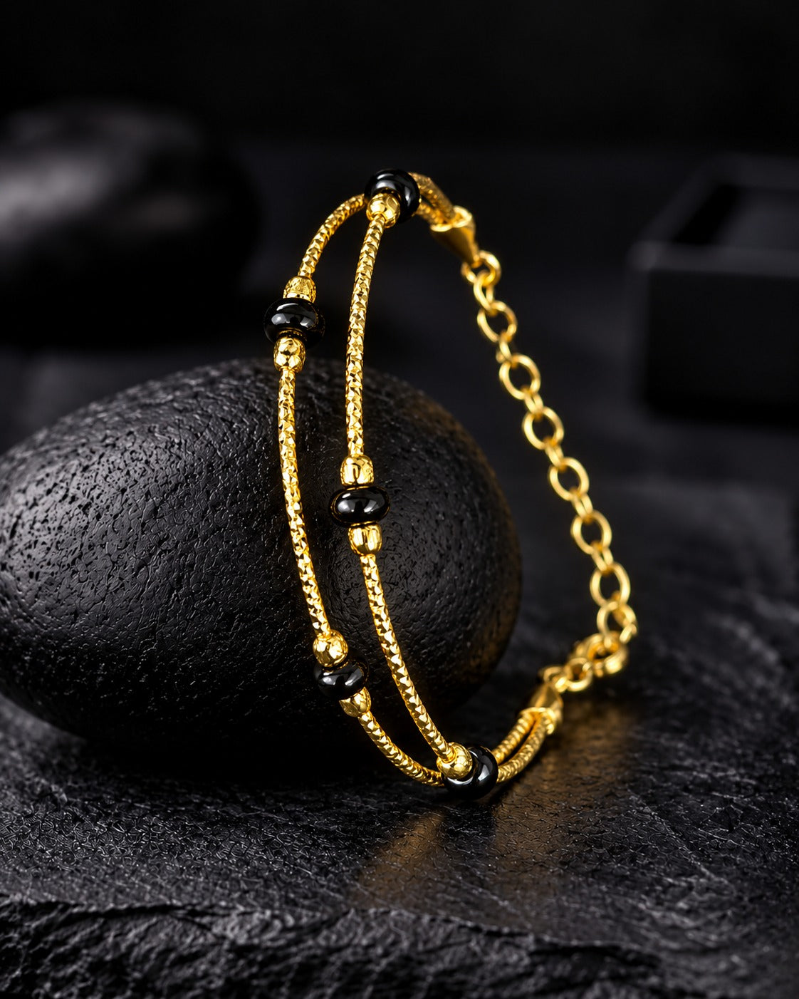 A gold-toned double-strand bracelet with black beads, draped over a dark textured stone on a dark surface.