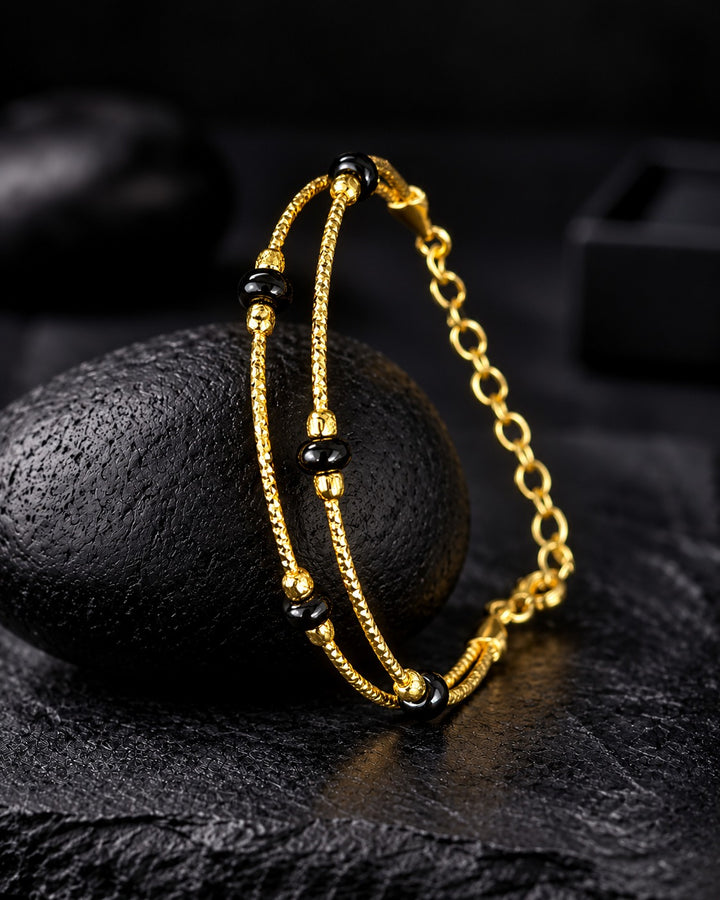 A gold-toned double-strand bracelet with black beads, draped over a dark textured stone on a dark surface.