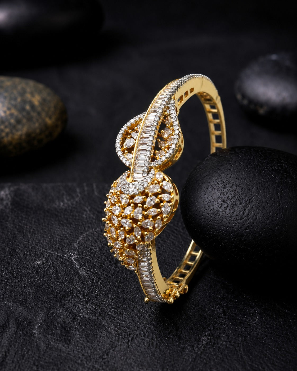 A gold-toned hinged bangle bracelet adorned with pavé-set clear stones, photographed on a dark textured surface with black stones in the background.