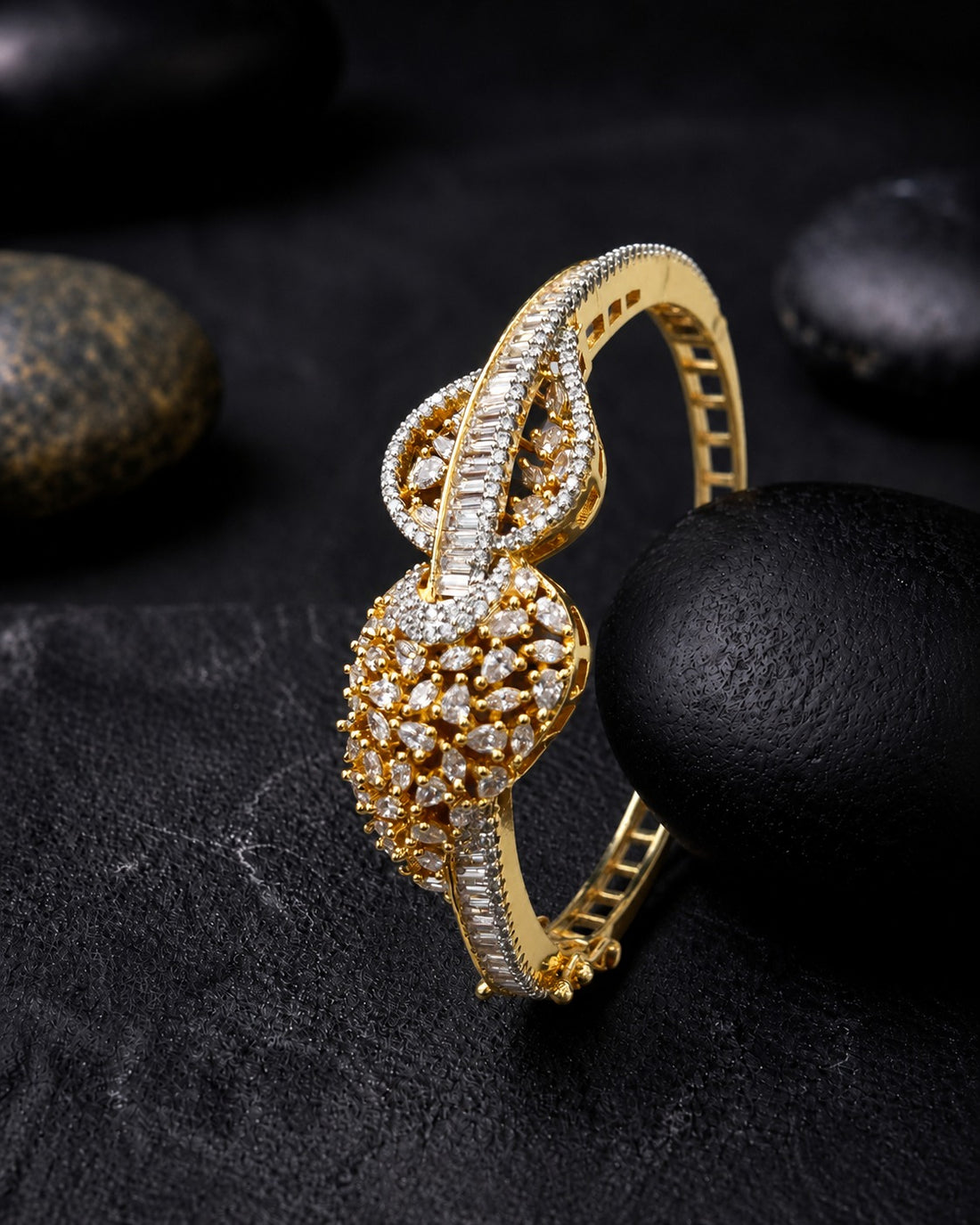A gold-toned hinged bangle bracelet adorned with pavé-set clear stones, photographed on a dark textured surface with black stones in the background.