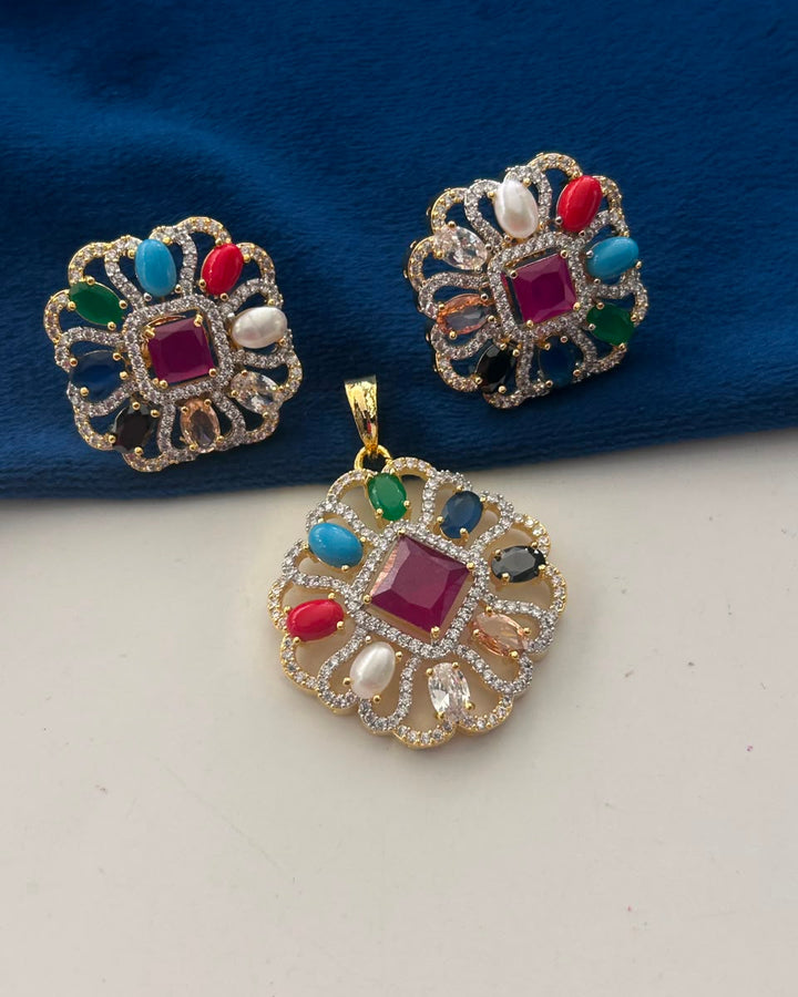 A gold-plated Indian style jewelry set featuring a matching pendant and earring with a central magenta square stone, surrounded by multicolored stones (red, blue, green, white) and clear pavé accents, displayed on a split white and blue background.