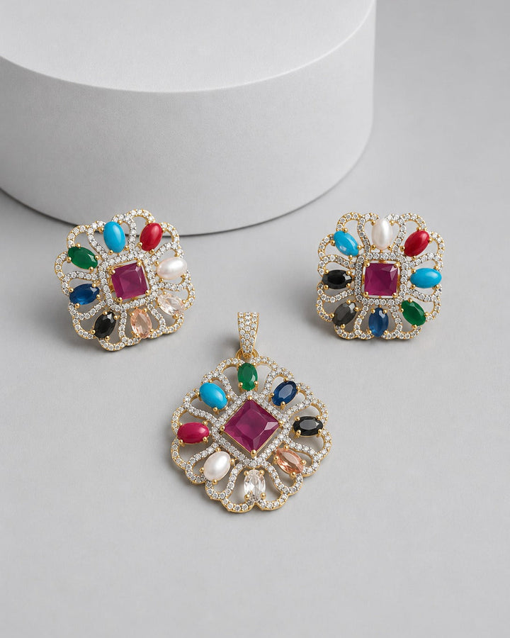 A gold-toned jewelry set featuring a central pendant and two matching square earrings, embellished with a colorful array of faux gemstones including pink, blue, green, red, black, and white, displayed on a light gray surface with a white cylindrical prop.