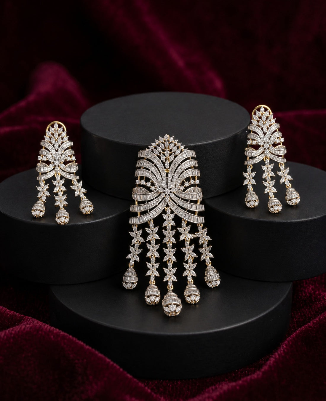 A gold-toned jewelry set featuring a large central pendent and two matching drop earrings, all adorned with clear stones, displayed on black cylindrical pedestals against a deep burgundy velvet background.