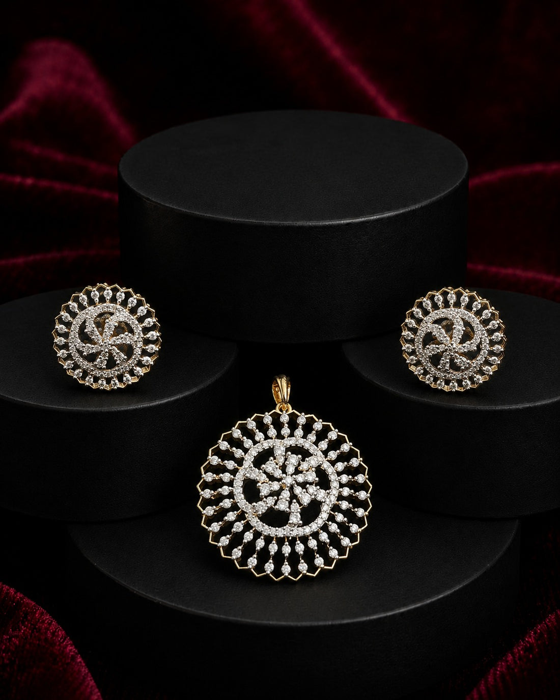A gold-toned jewelry set featuring a round pendant and matching stud earrings with intricate circular patterns set with clear stones, displayed on black cylindrical pedestals against a dark red velvet background.