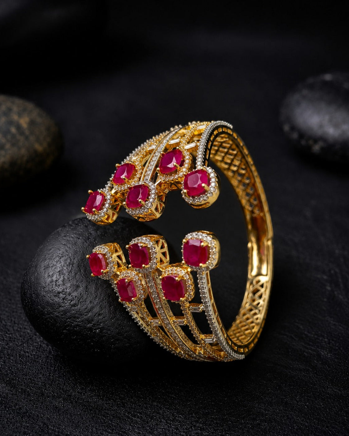 A gold-toned open cuff bracelet decorated with multiple round red gemstones and small clear stones, displayed on a dark textured surface with black stones in the background.