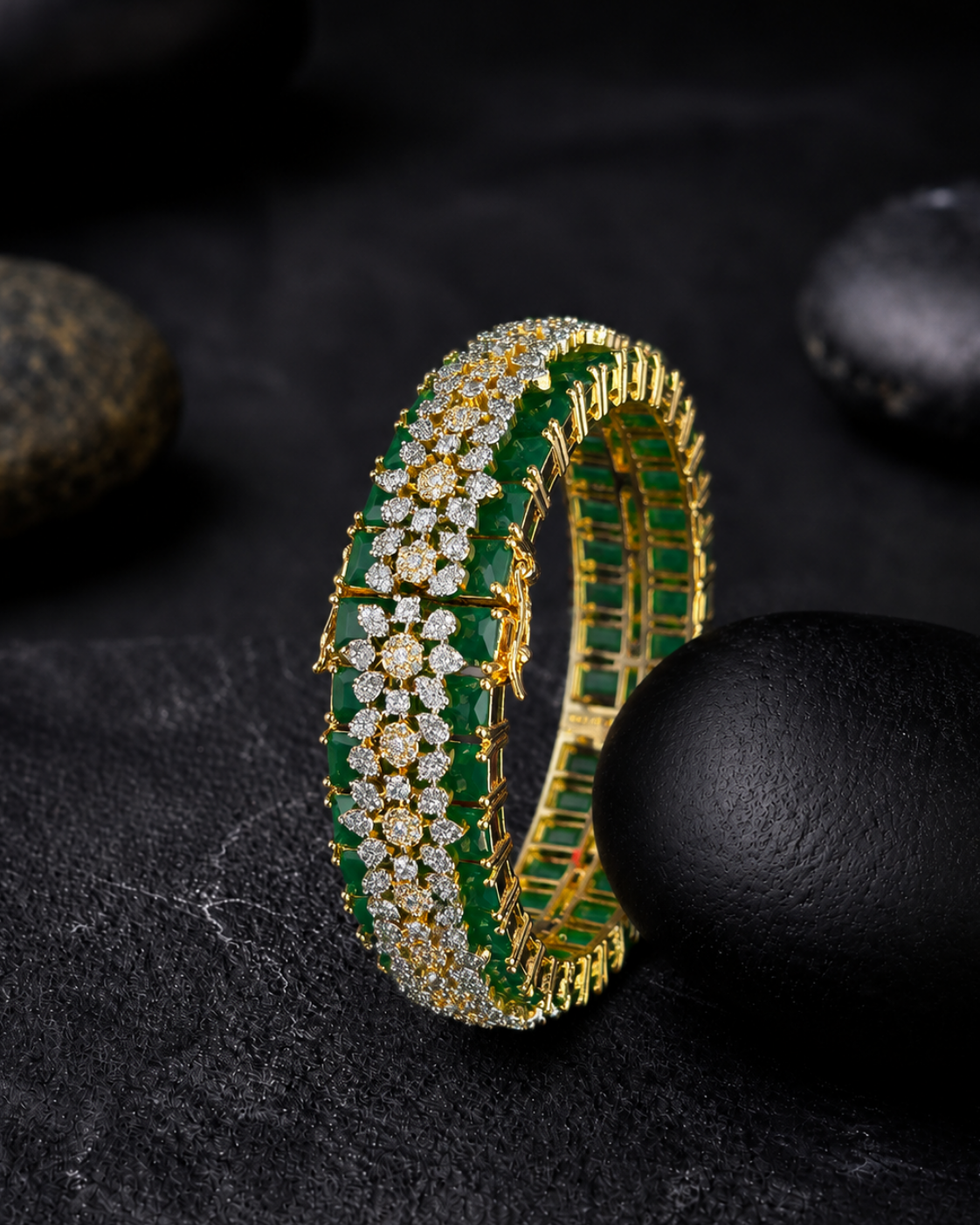 A gold-toned tennis-style bracelet with rows of green emerald-cut gemstones and clear stones, photographed on a dark textured surface with black stones around it.