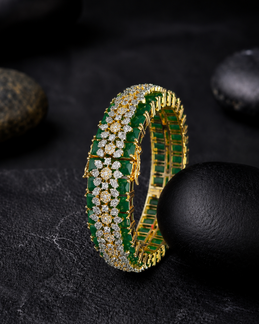 A gold-toned tennis-style bracelet with rows of green emerald-cut gemstones and clear stones, photographed on a dark textured surface with black stones around it.