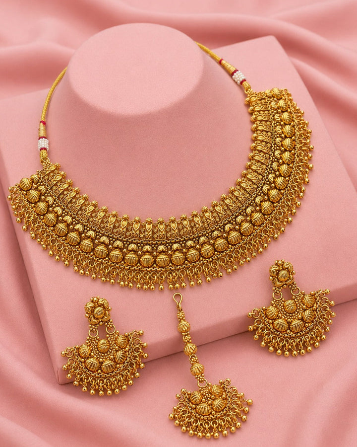 A gold-toned traditional jewelry set displayed on a pink mannequin stand and draped pink fabric. The set includes a wide filigree necklace, two matching dangling earrings, and a separate dangling charm.