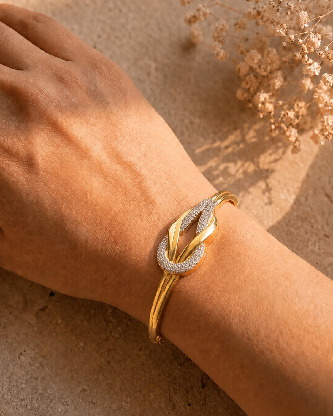 A gold-toned twist knot cuff bracelet with a textured, diamond-like pavé-set center, shown worn on a person's wrist against a warm beige textured background with dried flowers.