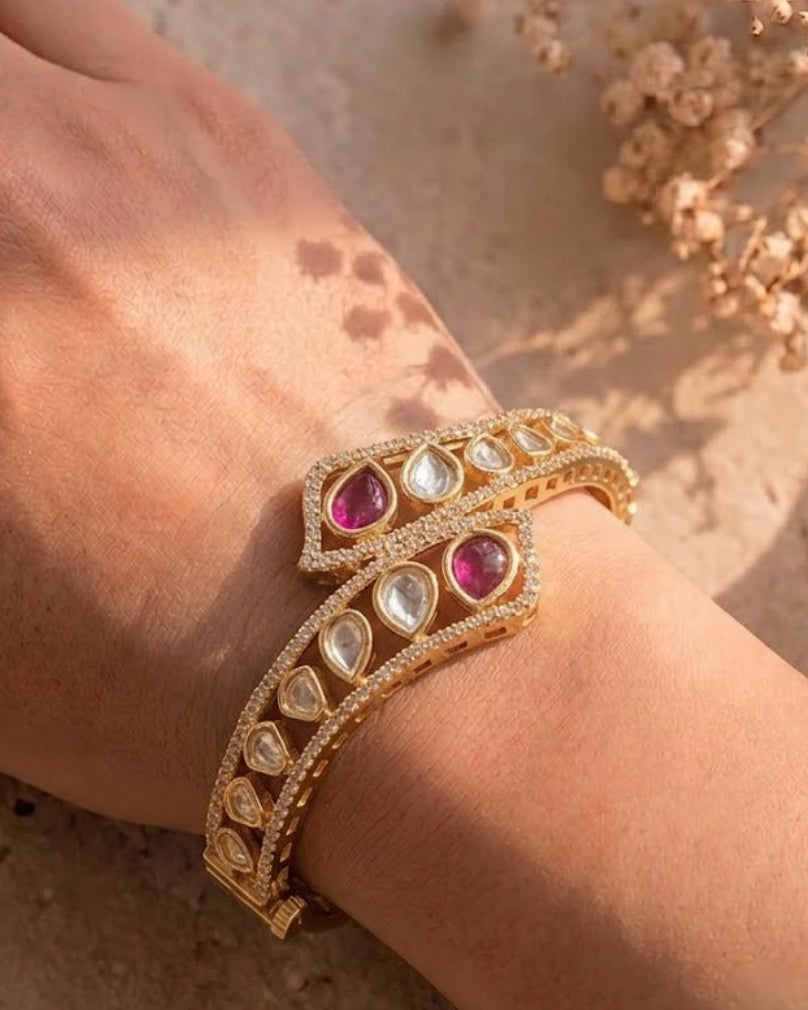 A gold-toned ornate bracelet with pink gemstones and clear stones, shown worn on a person's wrist against a warm beige textured background with dried flowers.