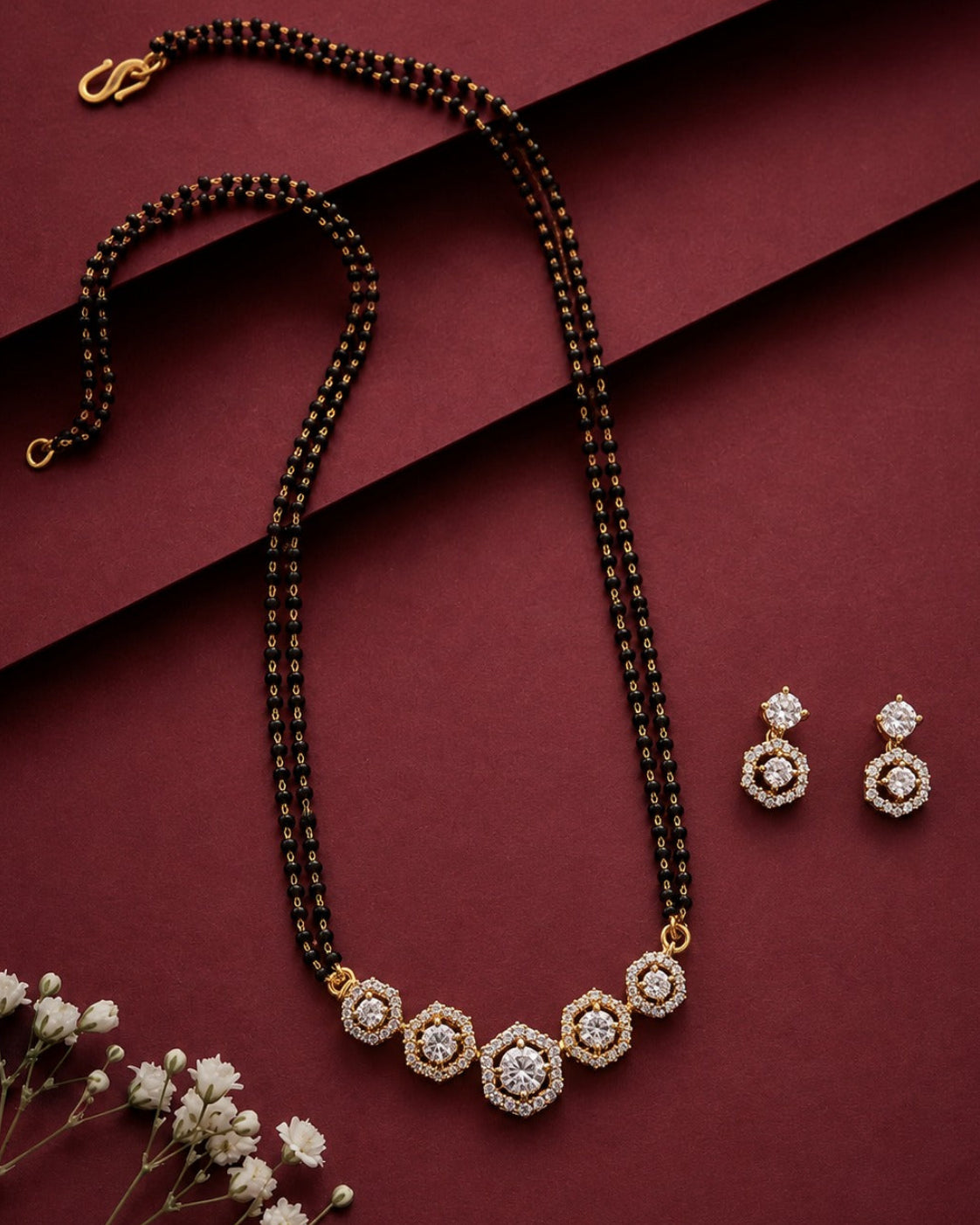 A gold and black beaded mangalsutra with a hexagonal crystal pendant, displayed alongside matching drop earrings on a dark burgundy background with decorative baby's breath flowers.