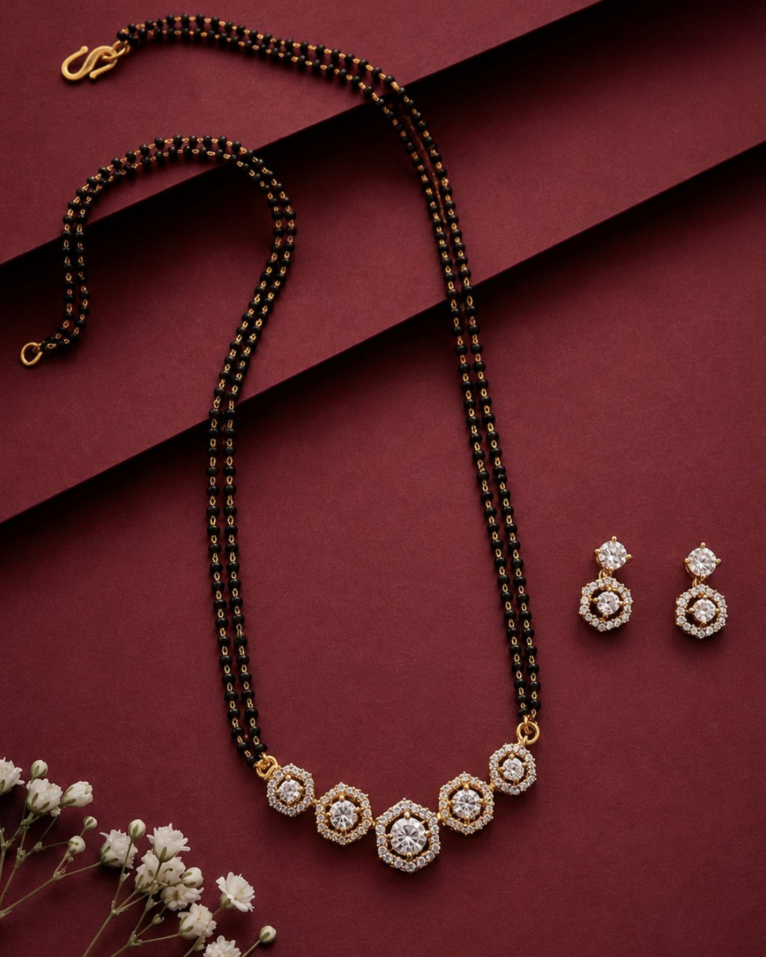 A gold and black beaded mangalsutra with a hexagonal crystal pendant, displayed alongside matching drop earrings on a dark burgundy background with decorative baby's breath flowers.