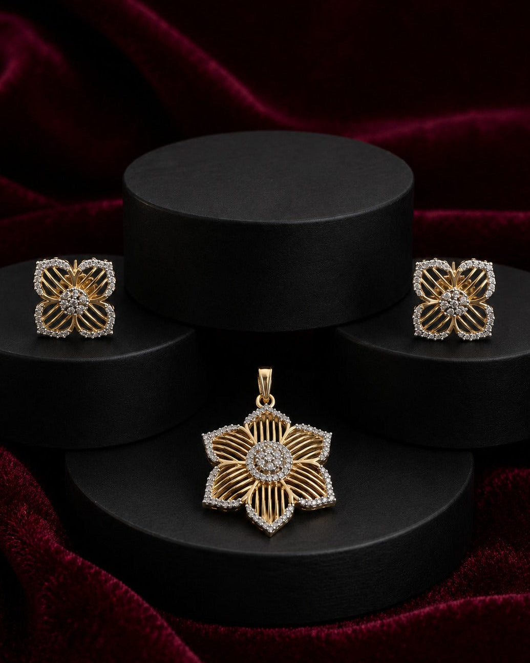 A gold and diamond jewelry set featuring a flower-shaped pendant, matching stud earrings, and a small matching charm, displayed on black cylindrical pedestals against a dark red velvet background.
