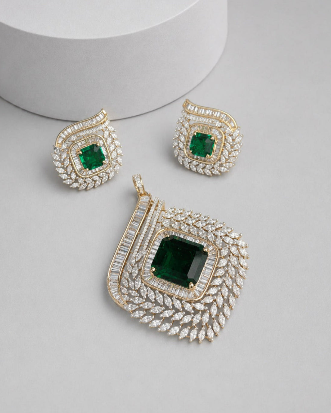 A gold and diamond jewelry set featuring a large pendant and matching earrings, each with a square-cut green emerald stone surrounded by pavé diamonds, displayed on a light gray surface.