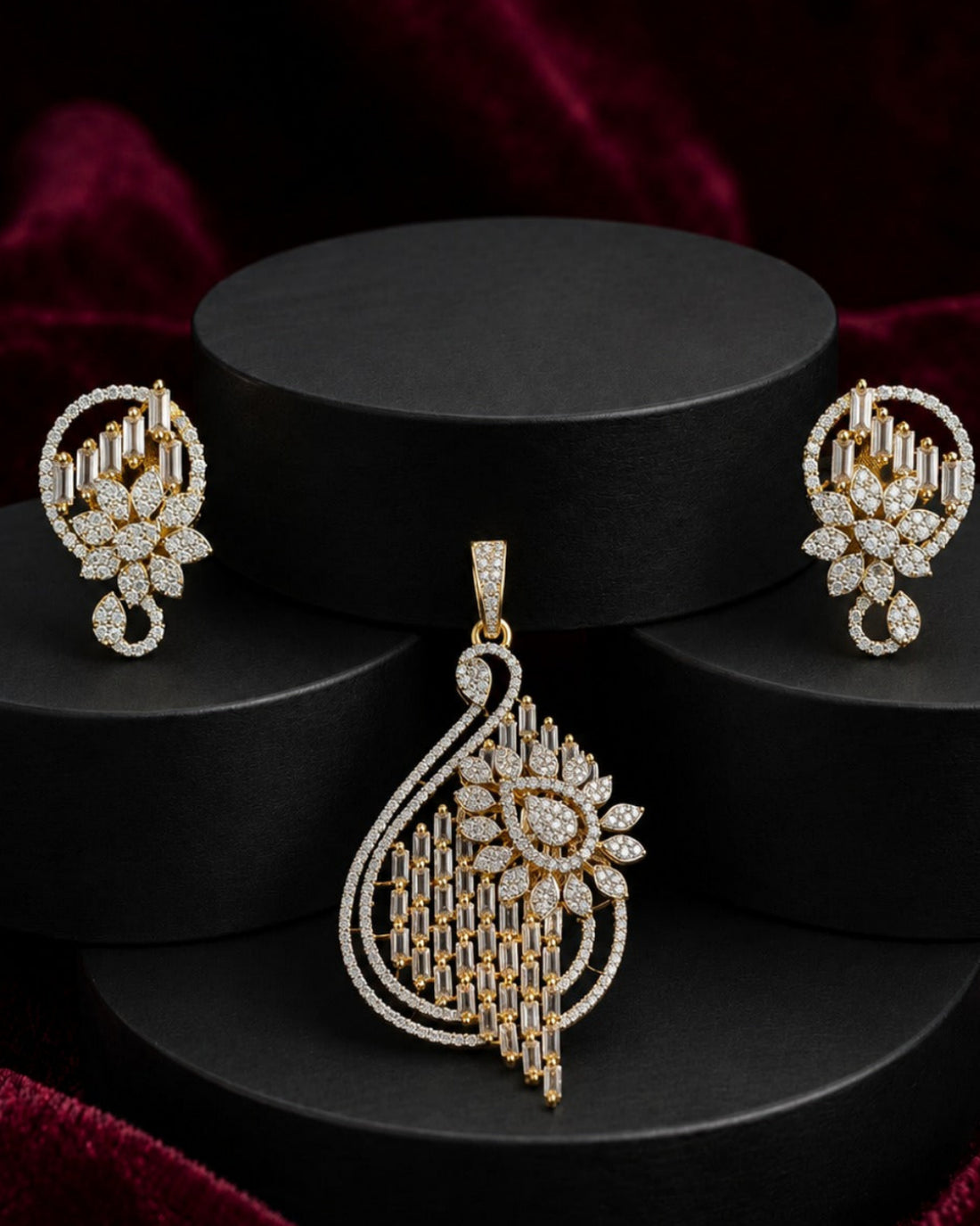 A gold and diamond jewelry set featuring a large teardrop pendant, matching earrings, and a small nose stud, displayed on black cylindrical pedestals against a dark red velvet background.