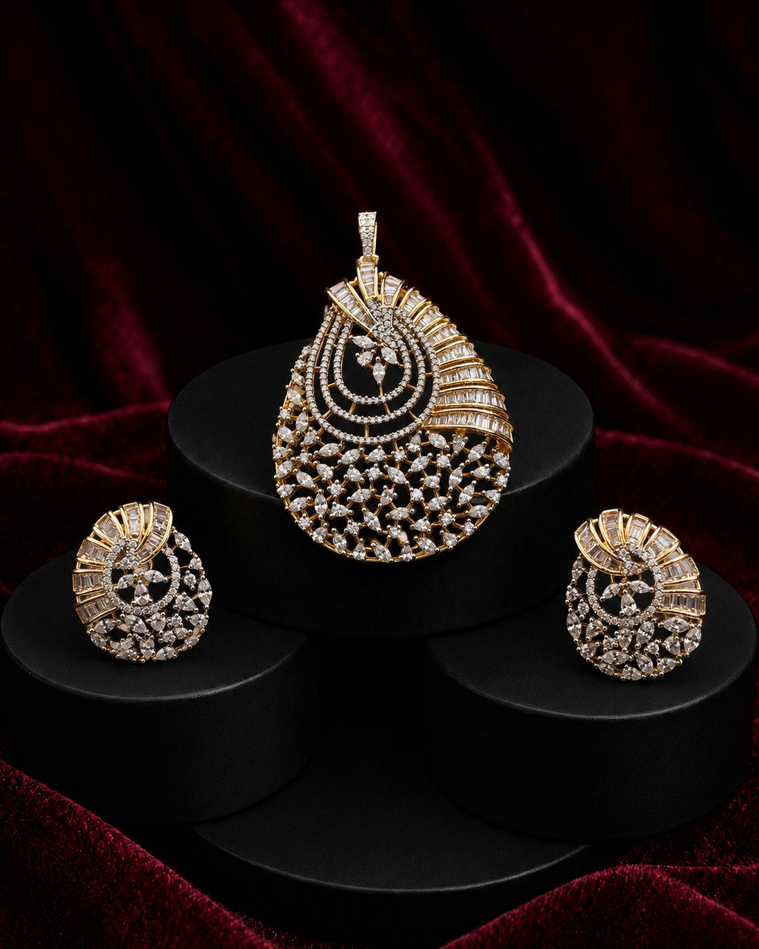 A gold and diamond jewelry set featuring a teardrop-shaped pendant, matching earrings, and a small nose stud, displayed on black cylindrical pedestals against a dark red velvet background.