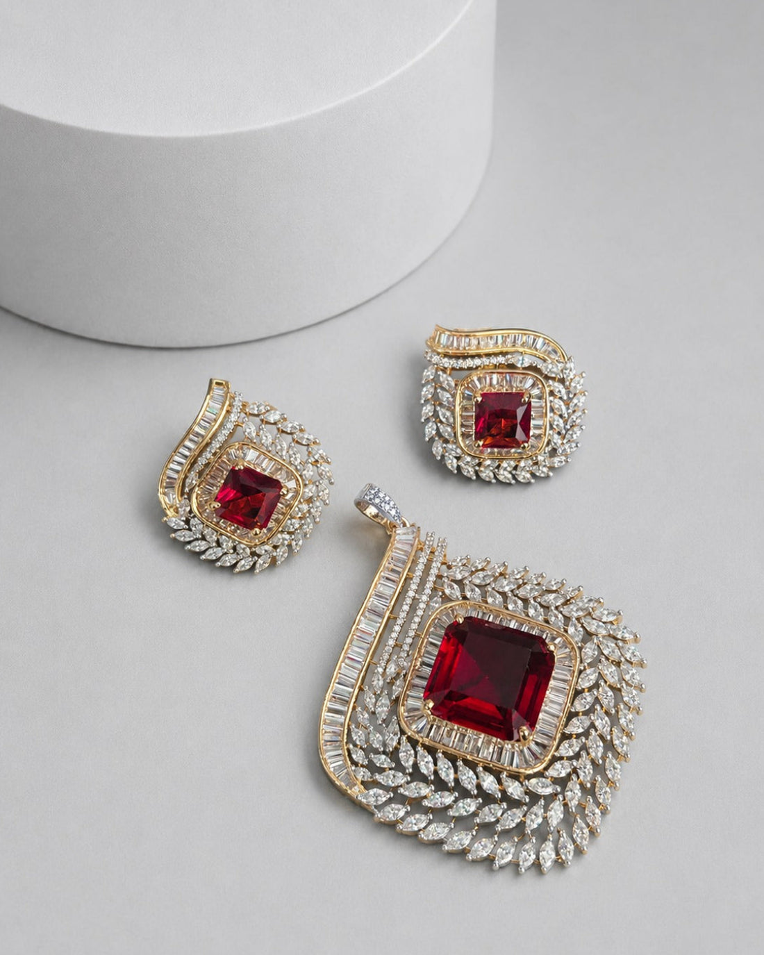 A gold and silver-toned jewelry set featuring a large pendant, matching earrings, and a small charm, each adorned with a central square red gemstone and surrounded by clear pavé stones, displayed on a light gray surface with a white cylindrical prop.
