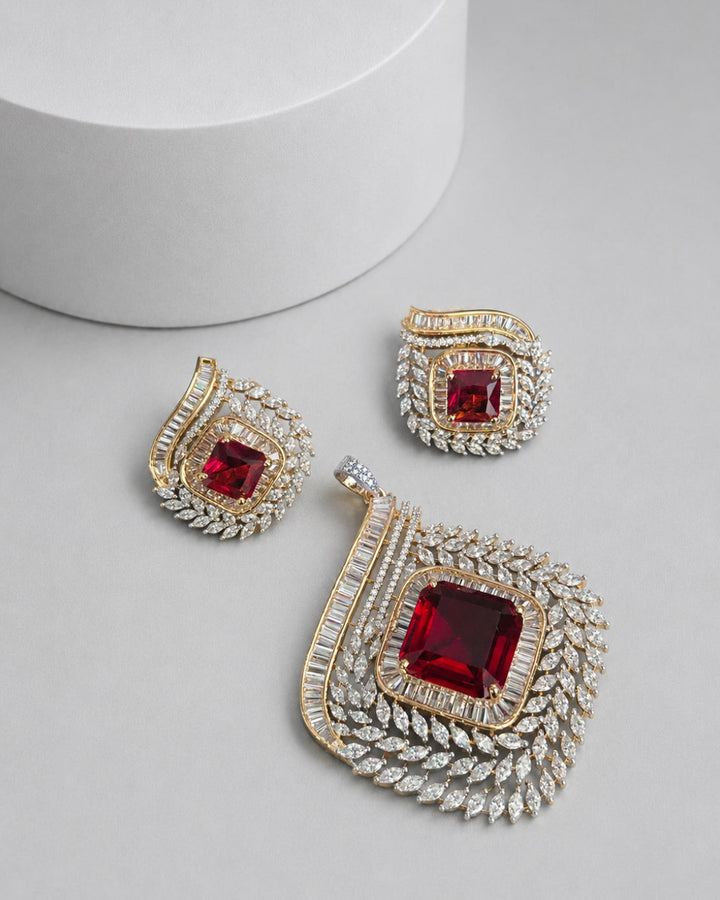 A gold and silver-toned jewelry set featuring a large pendant, matching earrings, and a small charm, each adorned with a central square red gemstone and surrounded by clear pavé stones, displayed on a light gray surface with a white cylindrical prop.