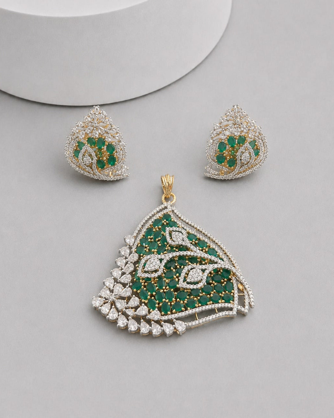 A gold and green stone jewelry set featuring a large teardrop-shaped pendant and matching stud earrings, displayed on a light gray surface with a white cylindrical prop.