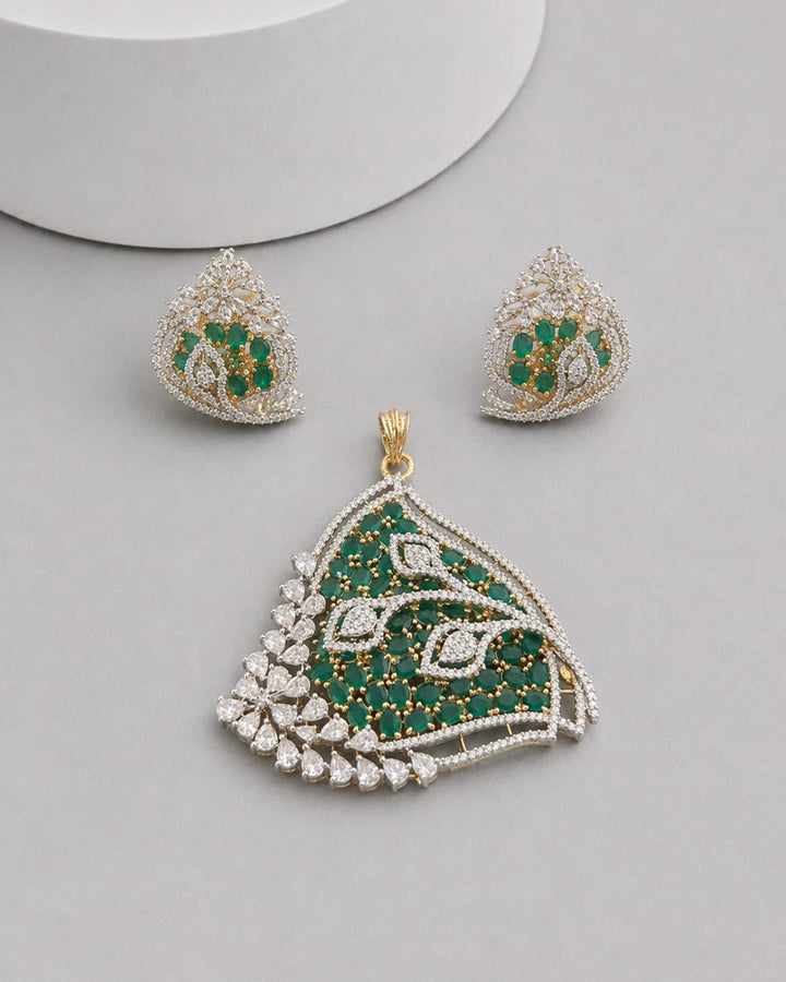 A gold and green stone jewelry set featuring a large teardrop-shaped pendant and matching stud earrings, displayed on a light gray surface with a white cylindrical prop.