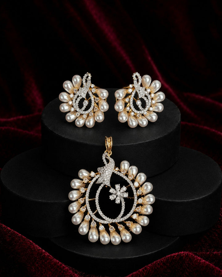 A gold and pearl jewelry set featuring a large peacock pendant, matching earrings, and a small necklace, displayed on black cylindrical pedestals against a dark red velvet background.