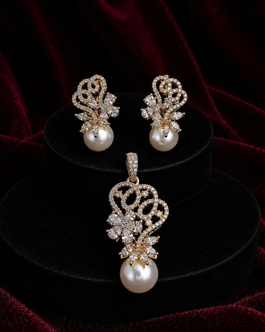 A gold and pearl jewelry set featuring a matching pendant and pair of stud earrings, displayed on black velvet risers against a dark red velvet background.