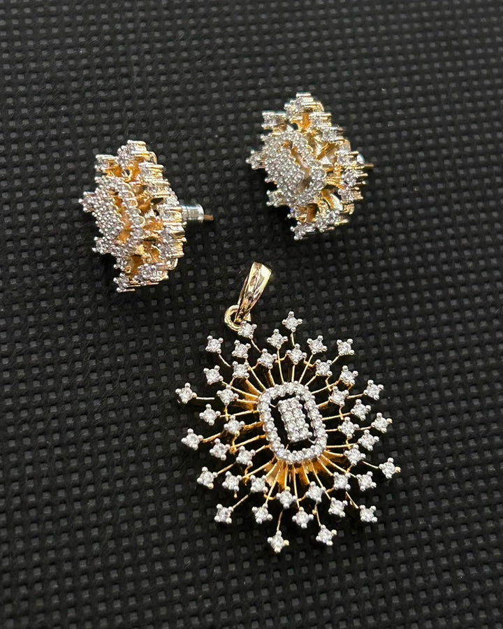 A gold and silver-toned jewelry set featuring a matching pendant and stud earring, both adorned with clear stones, displayed on a textured black background.
