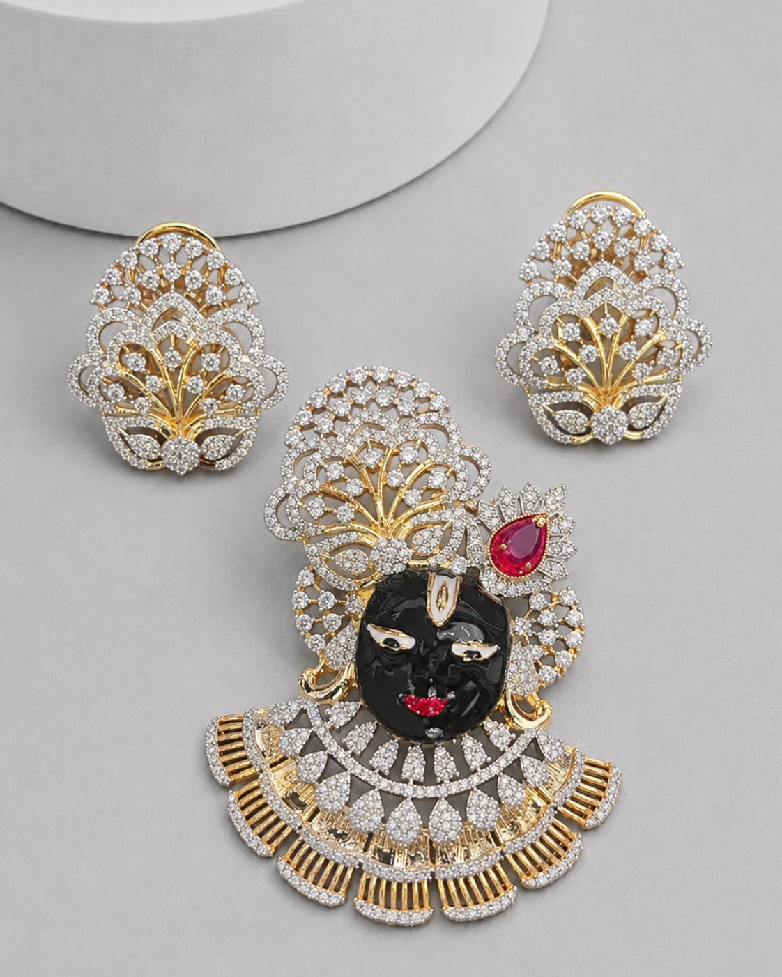 A gold and silver jewelry set featuring a central black and red deity pendant with a matching pair of stud earrings, all adorned with clear stones, displayed on a light gray surface.