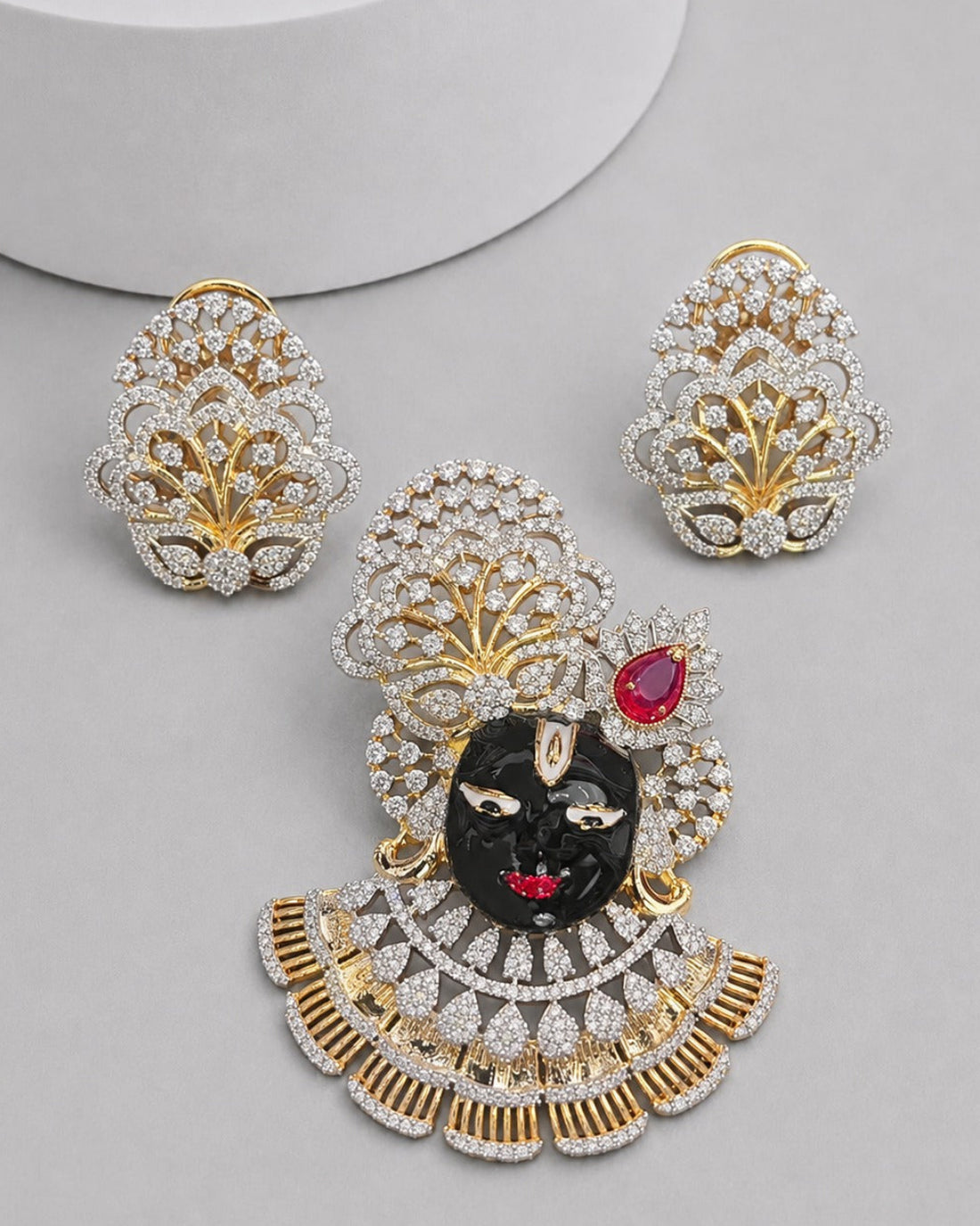 A gold and silver jewelry set featuring a central black and red deity pendant with a matching pair of stud earrings, all adorned with clear stones, displayed on a light gray surface.