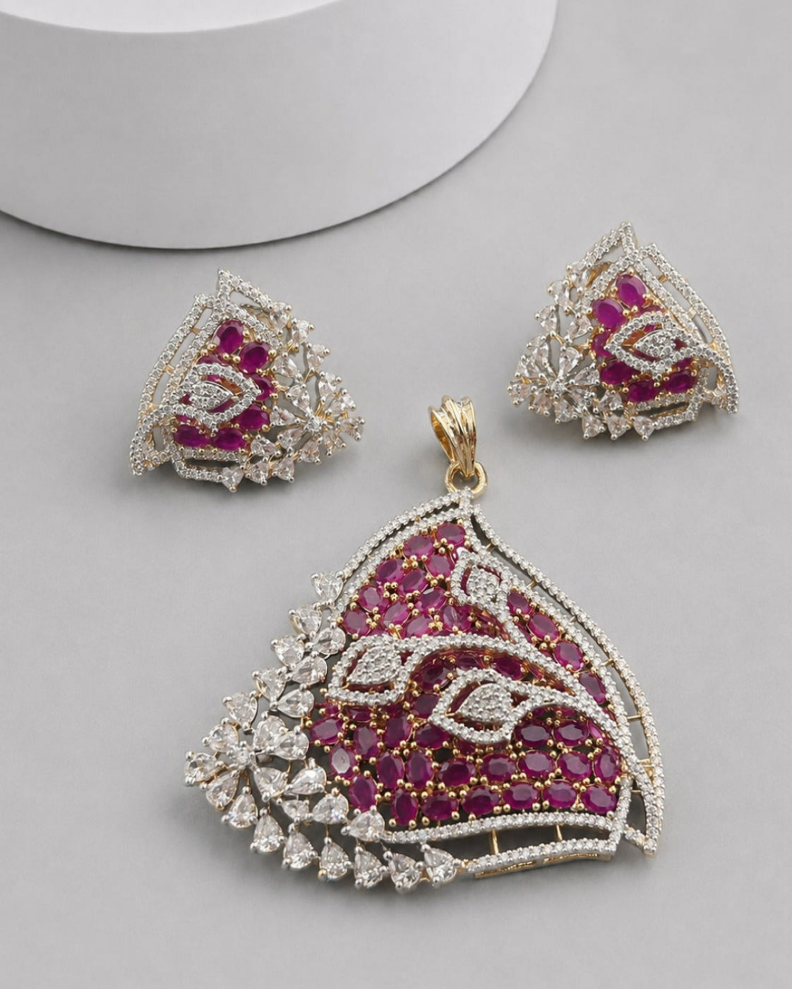A gold and silver jewelry set featuring a large scalloped pendant, matching triangle earrings, and a small box, all embellished with deep pinkish-red stones and clear crystals on a light gray background.