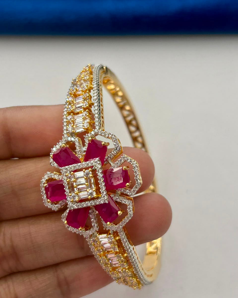 A gold and white gold bangle bracelet with a central floral motif featuring bright pink gemstones and clear baguette and round stones, held in a hand against a white background with a blue velvet surface in the background.