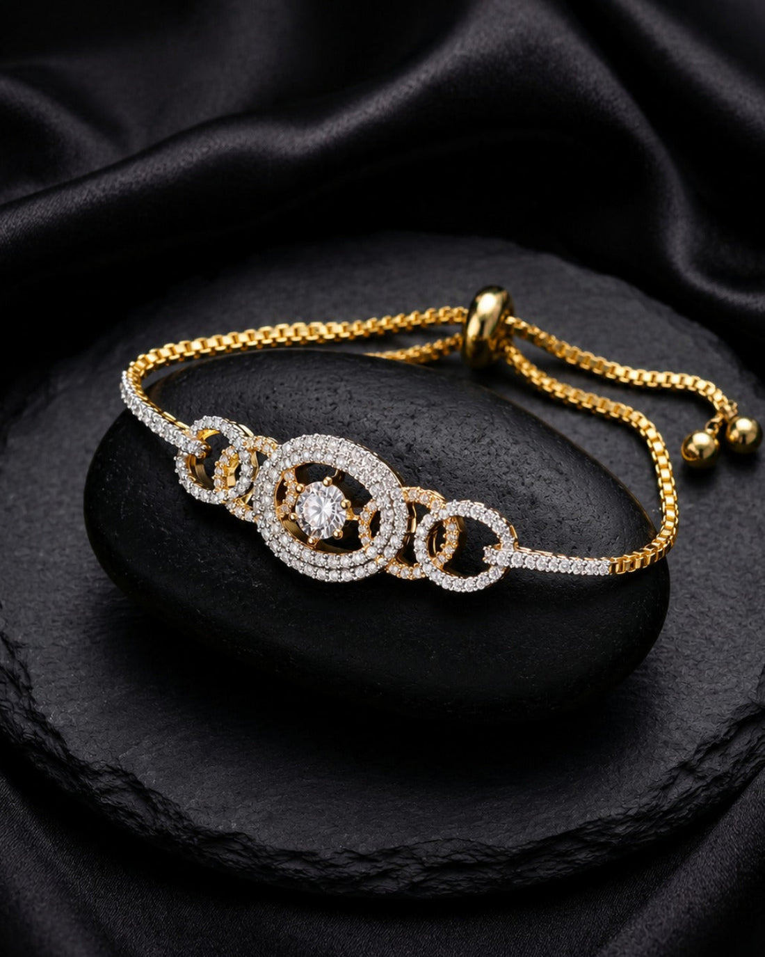 A gold and white stone bracelet with interlocking circular links, displayed on a black stone slab with draped black satin fabric in the background.