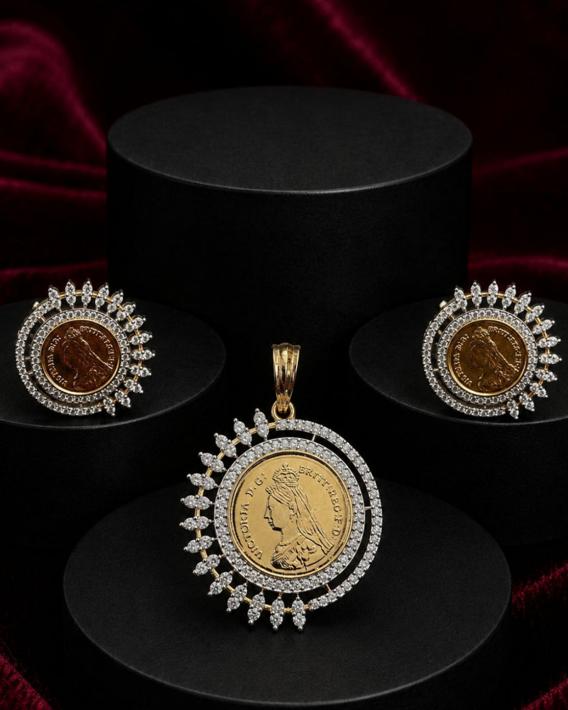 A gold and white stone jewelry set featuring a pendant and matching earrings, displayed on black cylindrical pedestals against a dark red velvet background. The pendant and earrings feature a central gold coin surrounded by a halo of sparkling white stones.