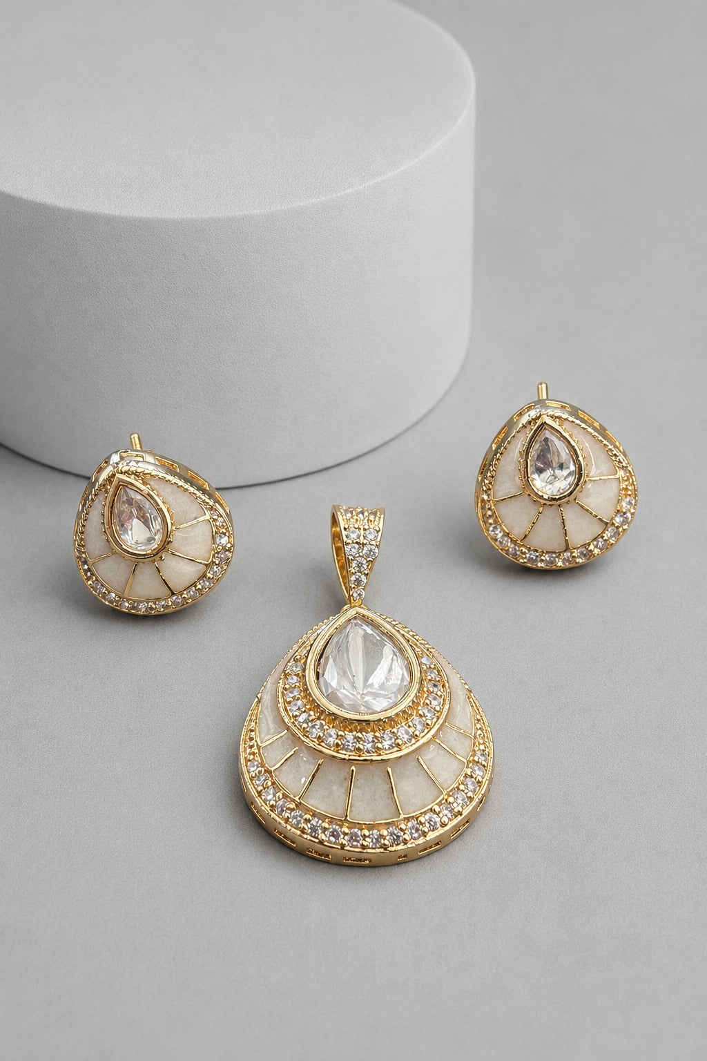 A gold and white teardrop-shaped jewelry set featuring a pendant, matching earrings, and small stud earrings, displayed on a gray surface with a white cylindrical prop.