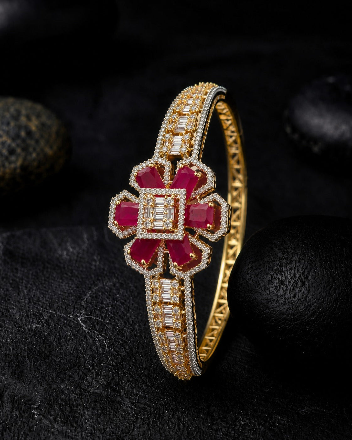 A gold bangle bracelet featuring a central floral motif with deep red gemstones and rows of clear stones, displayed on a dark textured surface with black stones in the background.
