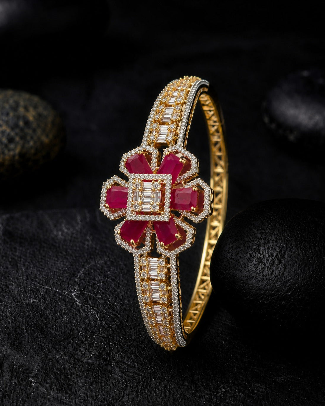 A gold bangle bracelet featuring a central floral motif with deep red gemstones and rows of clear stones, displayed on a dark textured surface with black stones in the background.