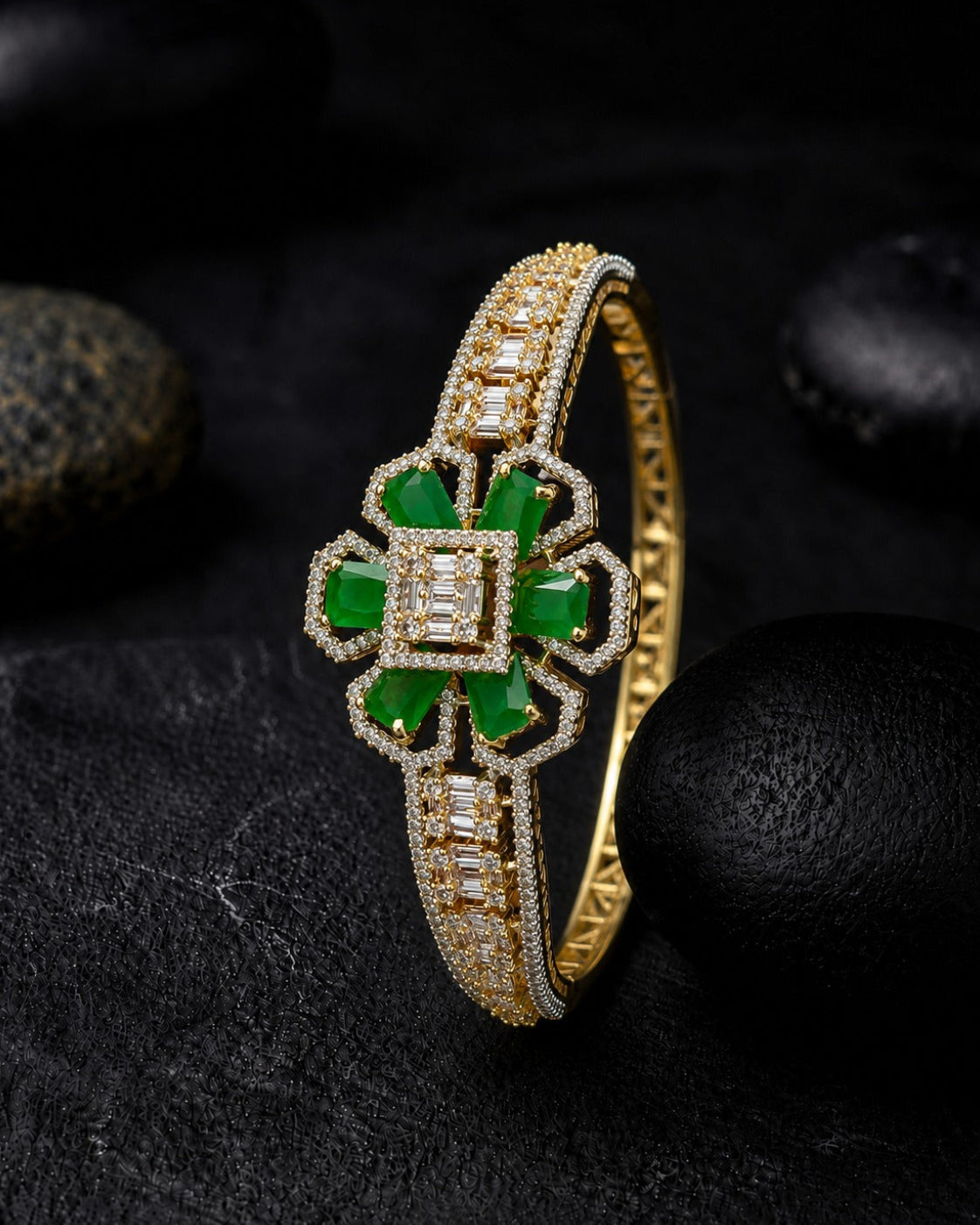 A gold bangle bracelet featuring a central four-leaf clover motif with vibrant green stones and diamond-like clear accents, displayed on a dark textured surface with black stones.