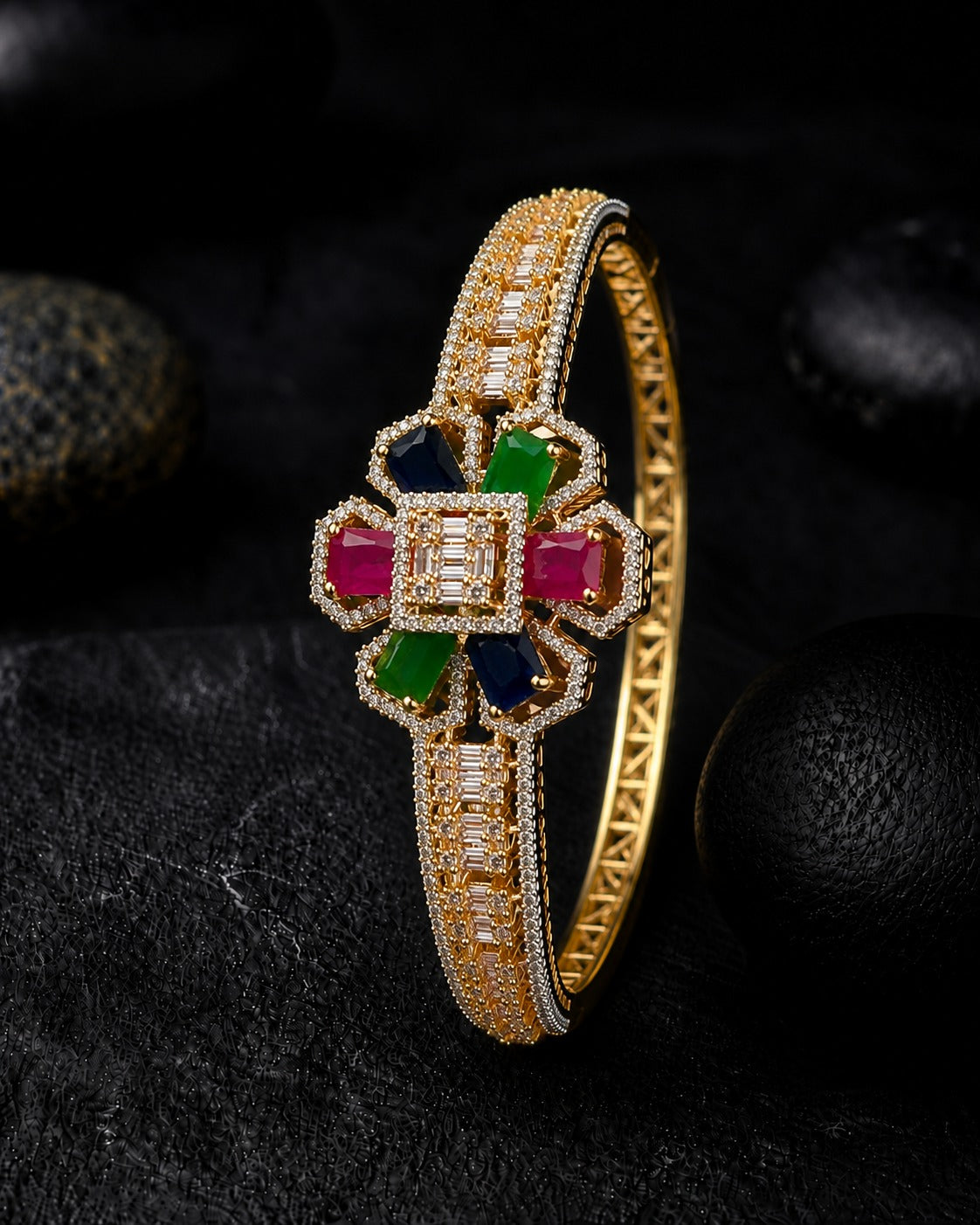 A gold bangle bracelet with a floral centerpiece featuring rectangular pink, green, and blue gemstones, surrounded by clear pavé stones, set against a dark textured background with black stones.