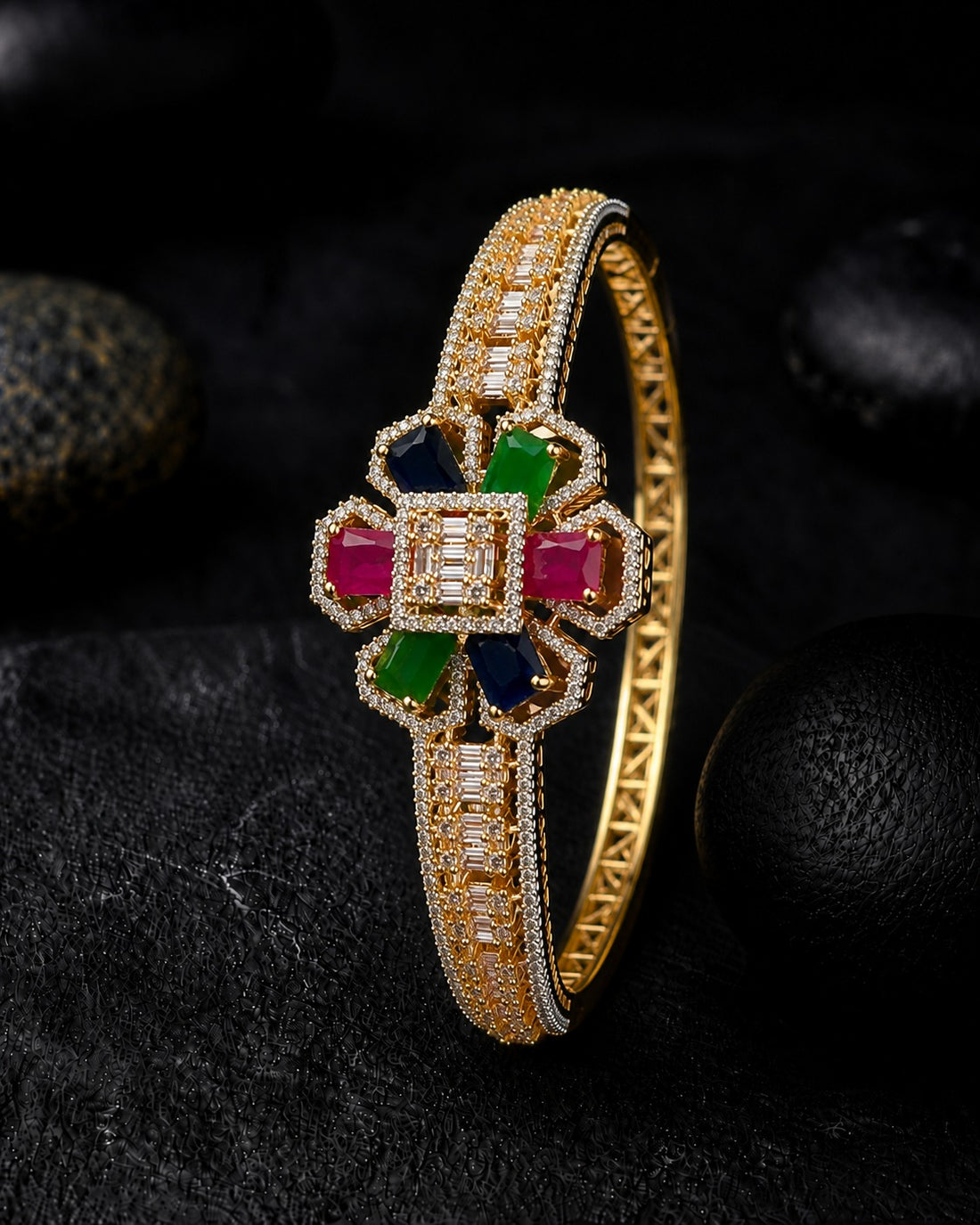 A gold bangle bracelet with a floral centerpiece featuring rectangular pink, green, and blue gemstones, surrounded by clear pavé stones, set against a dark textured background with black stones.