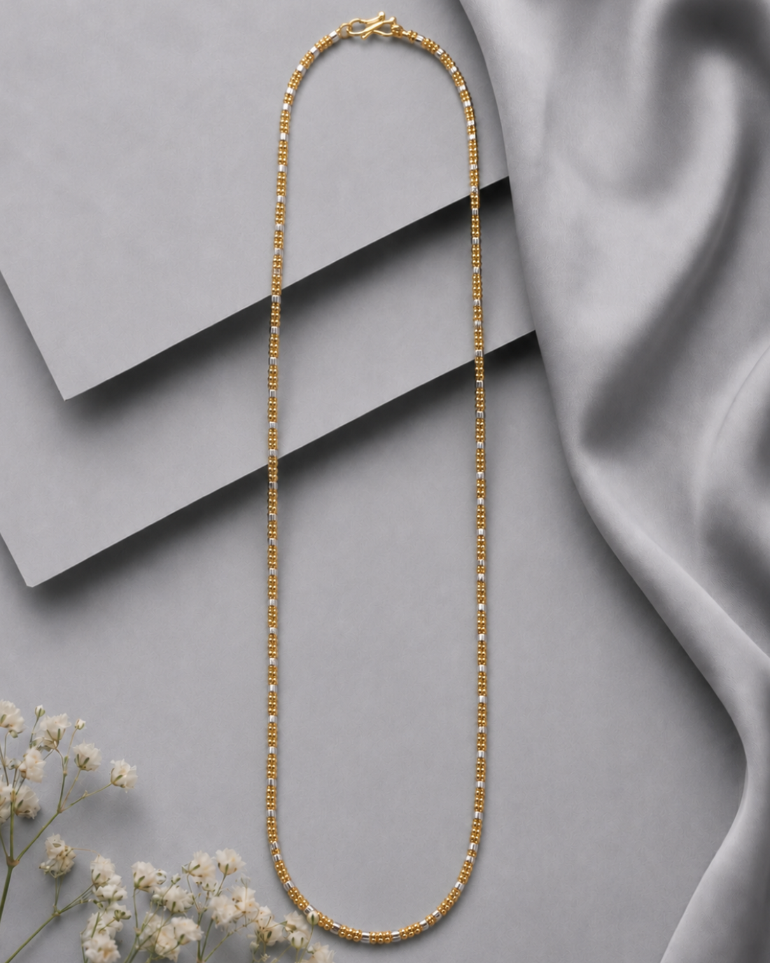 A gold beaded chain necklace arranged in a long oval shape on a gray background with layered geometric blocks and draped satin fabric, with small white flowers in the lower left corner.