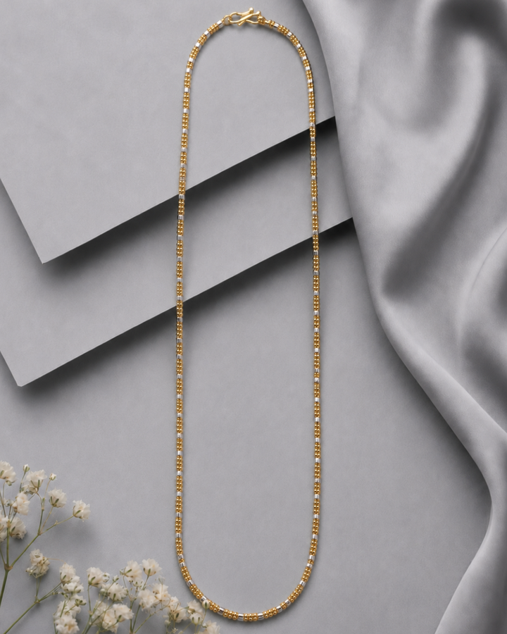A gold beaded chain necklace arranged in a long oval shape on a gray background with layered geometric blocks and draped satin fabric, with small white flowers in the lower left corner.