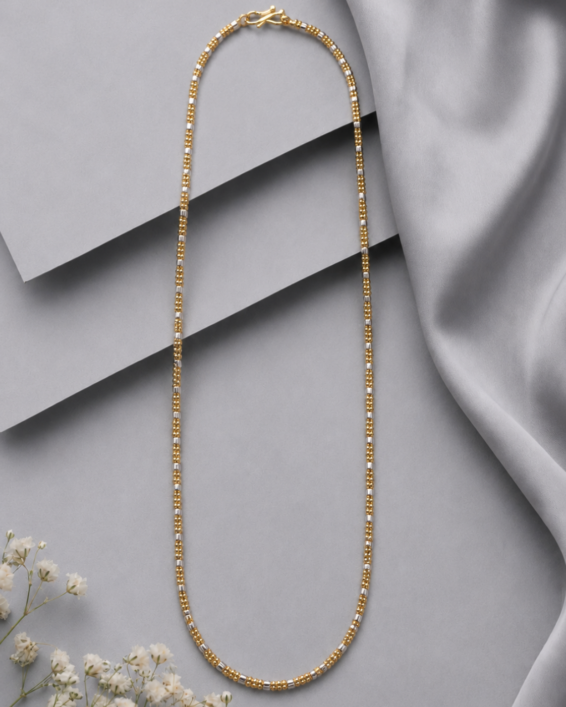 A gold beaded chain necklace arranged in an oval shape on a gray background with layered geometric blocks and draped satin fabric, with small white flowers in the lower left corner.
