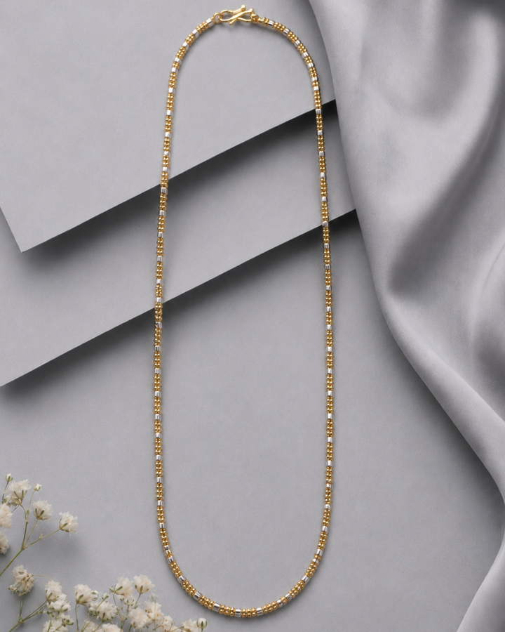 A gold beaded chain necklace arranged in an oval shape on a gray background with layered geometric blocks and draped satin fabric, with small white flowers in the lower left corner.