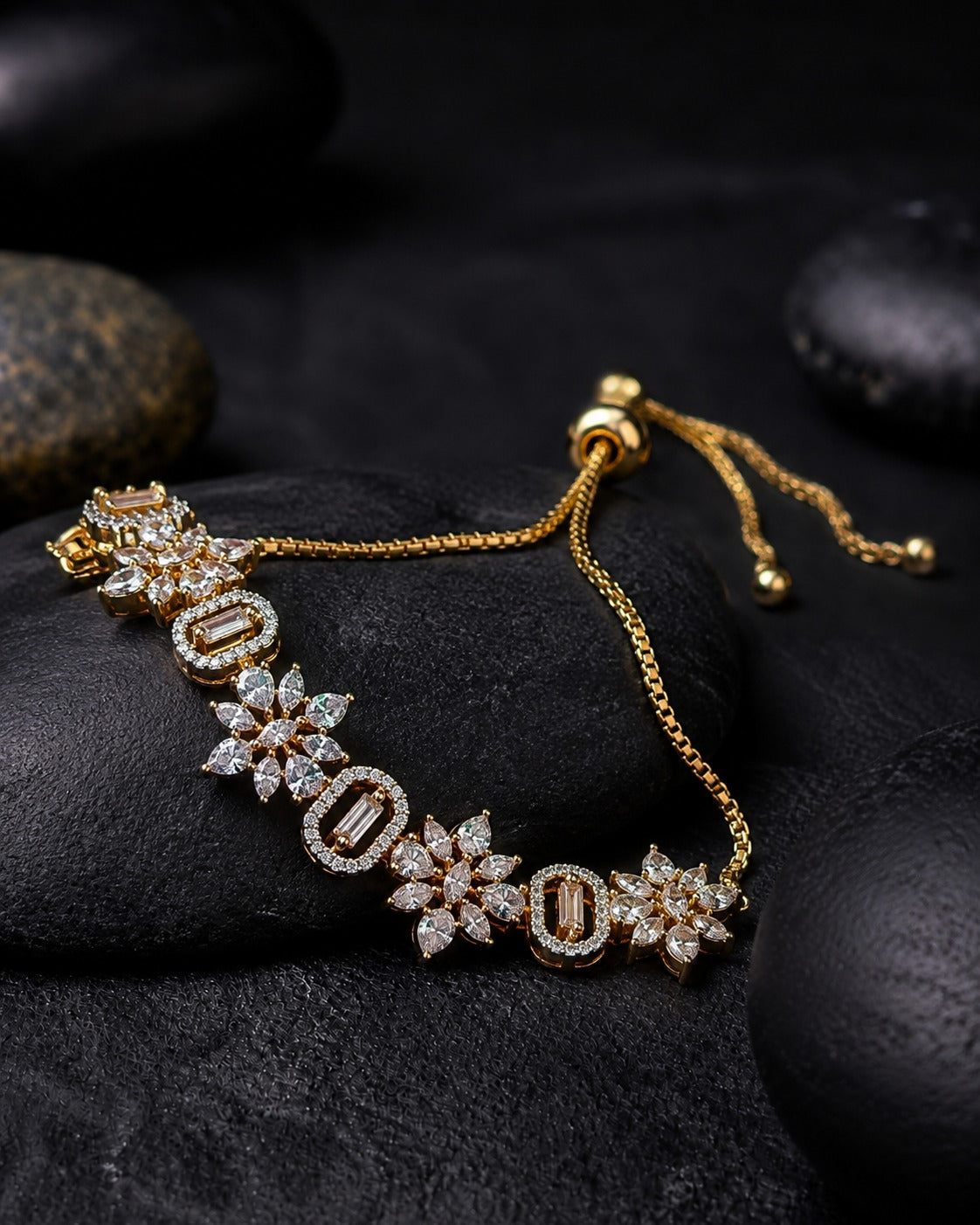 A gold chain bracelet with alternating oval and floral emerald-cut stone motifs, displayed on dark textured stones.