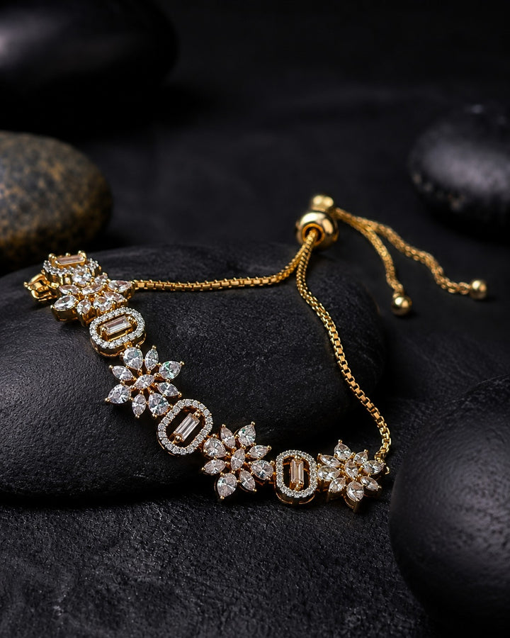 A gold chain bracelet with alternating oval and floral emerald-cut stone motifs, displayed on dark textured stones.