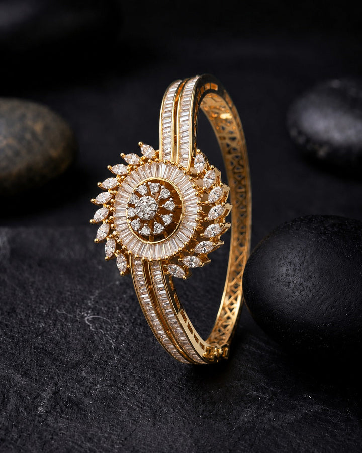 A gold hinged bangle bracelet decorated with a circular sunburst motif set with clear stones, accented by three parallel rows of channel-set stones, displayed on a dark textured surface with black stones.