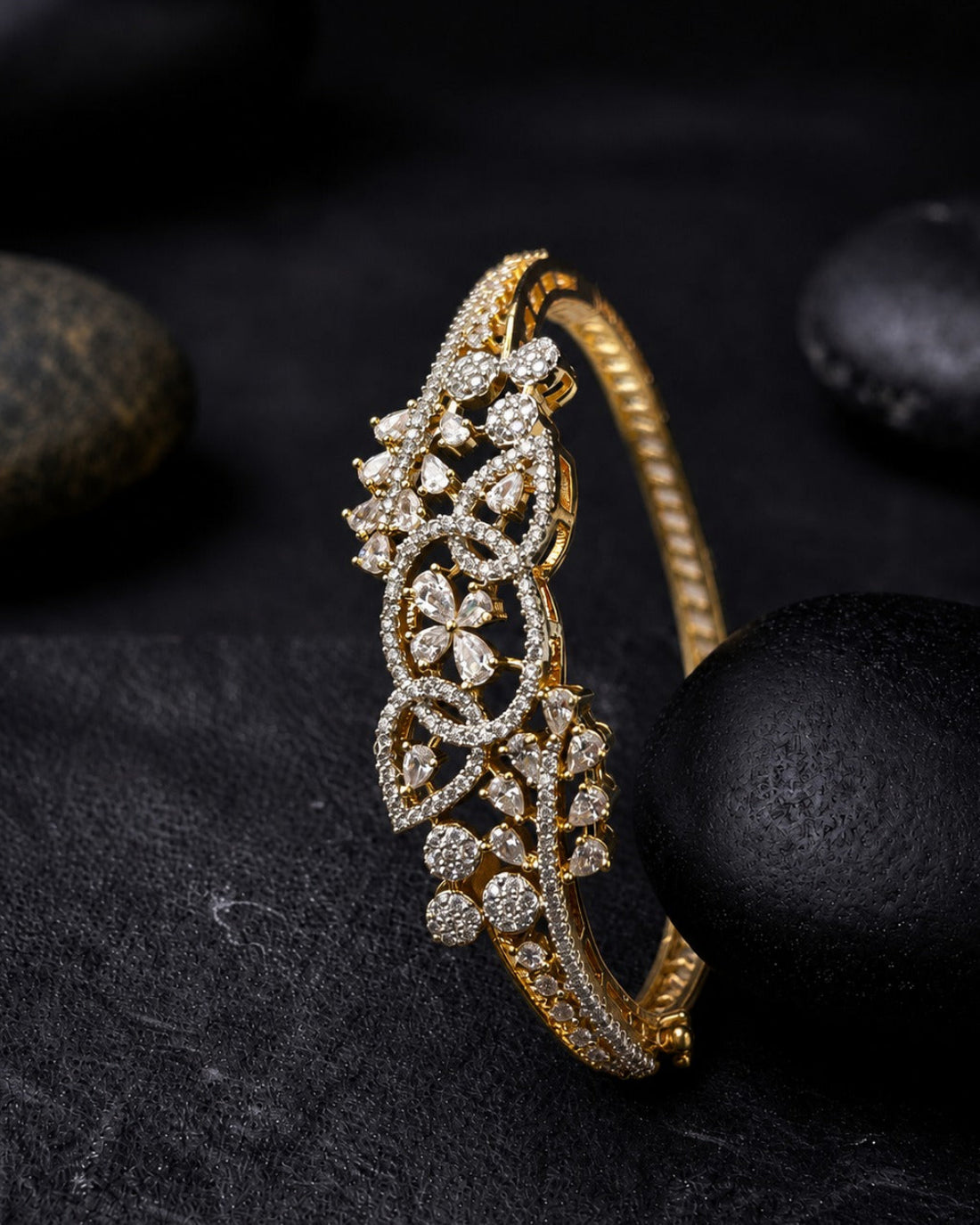 A gold hinged bangle bracelet decorated with intricate pavé diamond floral motifs, displayed on a dark textured surface with black stones.