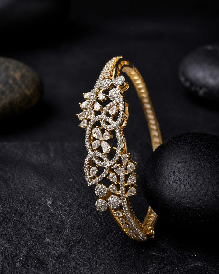 A gold hinged bangle bracelet decorated with intricate pavé diamond floral motifs, displayed on a dark textured surface with black stones.
