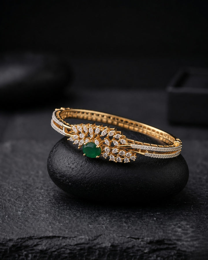 A gold hinged bangle bracelet with a central green emerald-cut stone and surrounding pavé-set clear stones, displayed on a smooth black stone against a dark textured background.