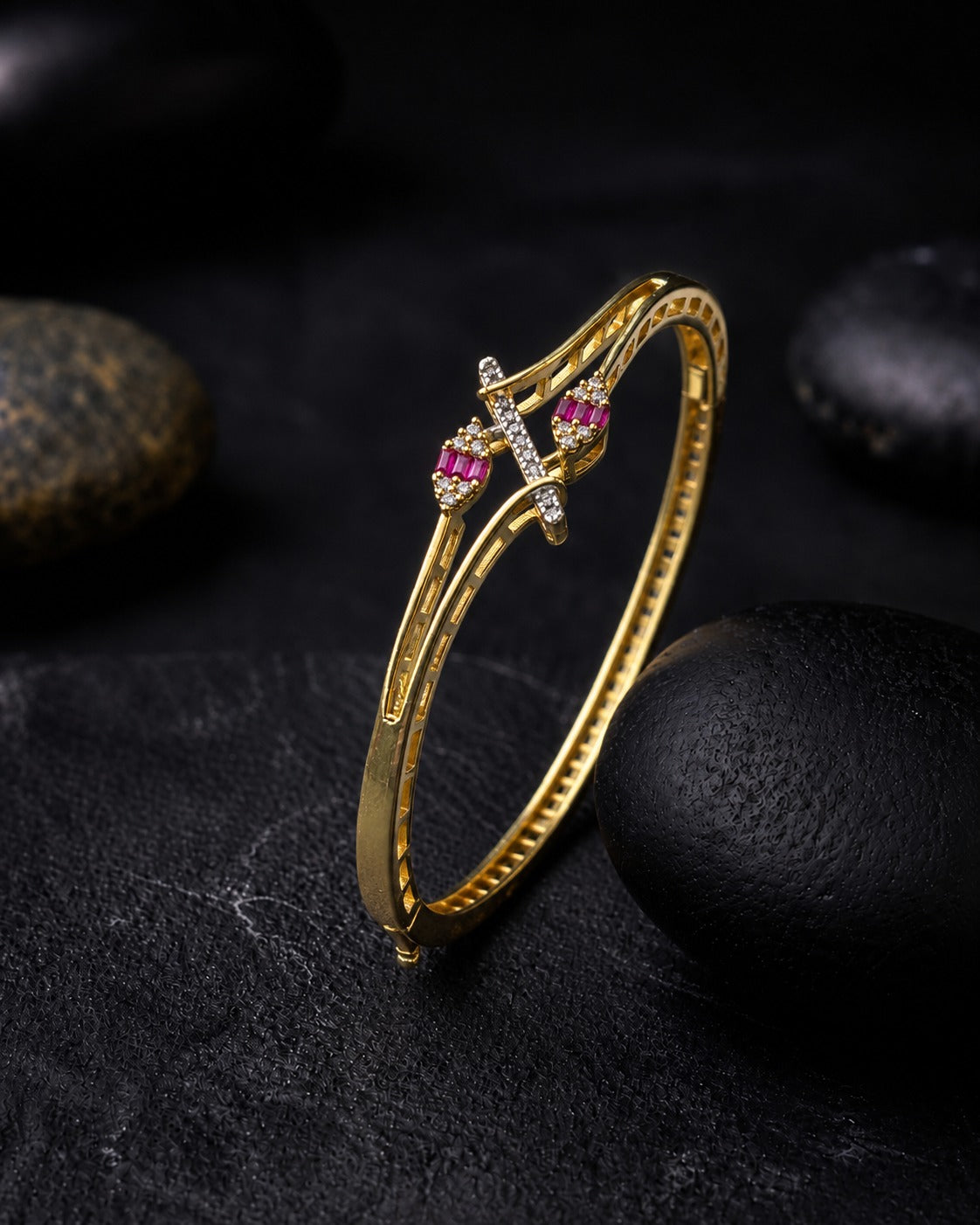 A gold hinged bangle bracelet with a decorative cross-shaped center featuring pink gemstones and small clear stones, displayed on a dark textured surface with black stones.